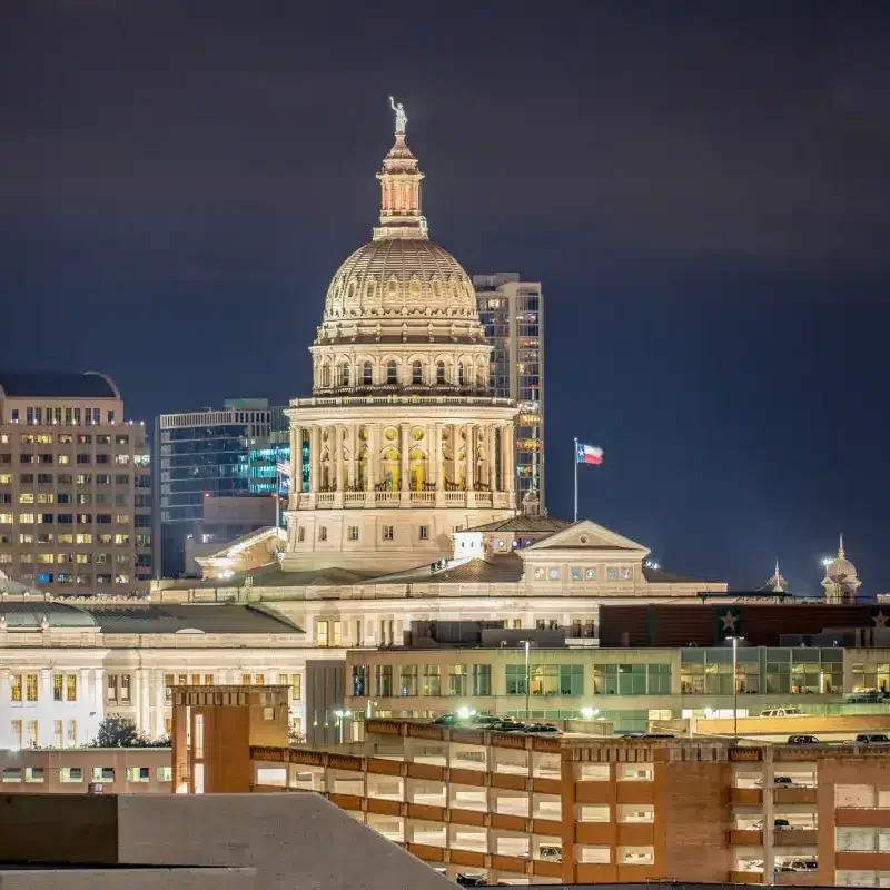 Brightly lit Texas Capitol at nighttime with city lights behind it.