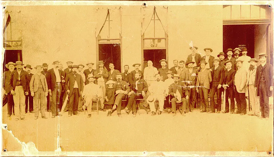 Seated governor and high officials with standing employees, mostly men in hats, outside a building with four doors. 