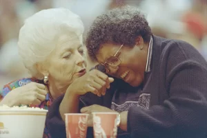 Barbara Jordan laughing while Governor Ann Richards speaks in her ear. A bucket of popcorn and two cokes are between them.