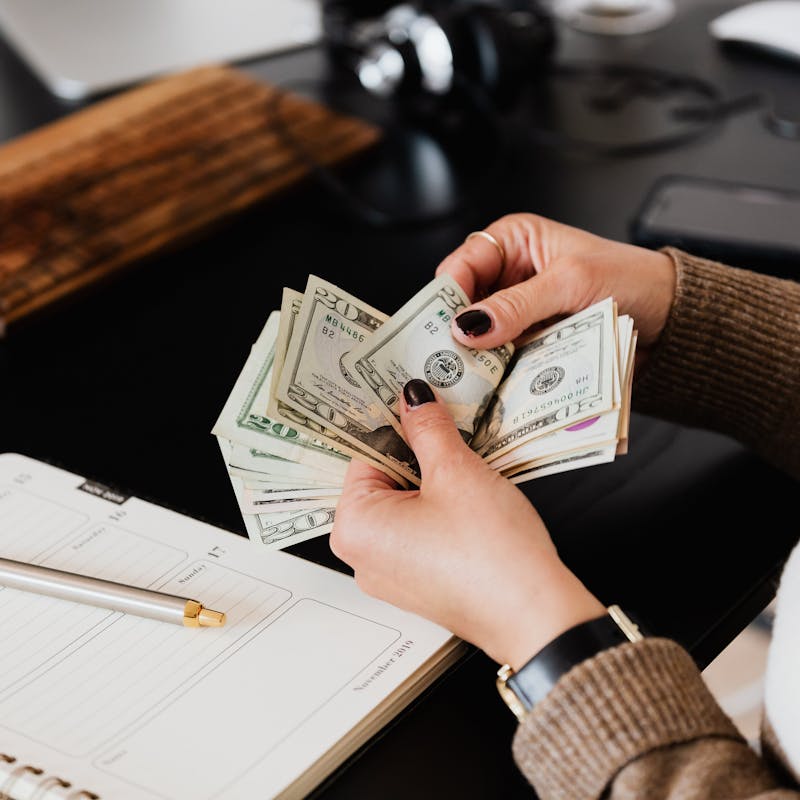 Unrecognizable elegant female in sweater counting dollar bills while sitting at wooden table with planner and pen